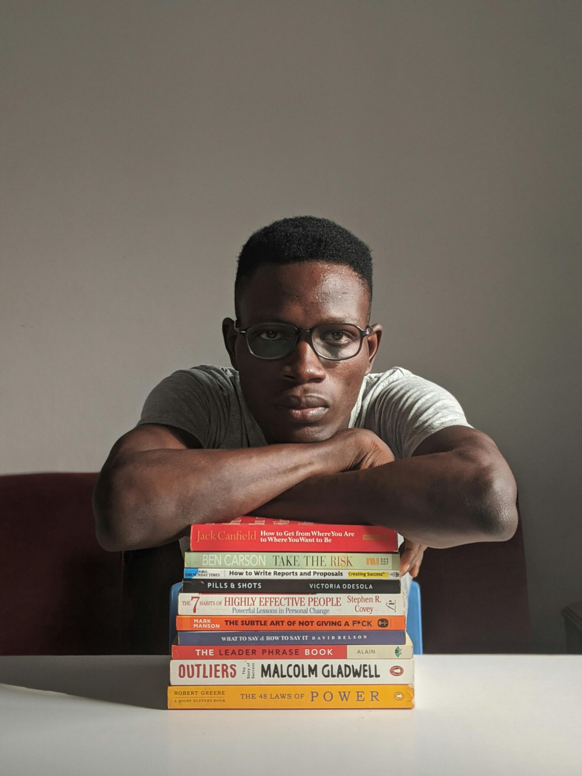 A man with eyeglasses sitting indoors, leaning on a stack of books with a serious expression.